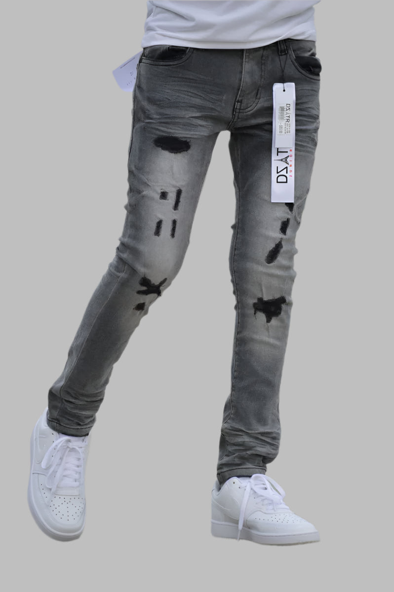 A pair of gray DZTR ripped stretch jeans with multiple rips, displayed on a model wearing white sneakers and a light-colored top.