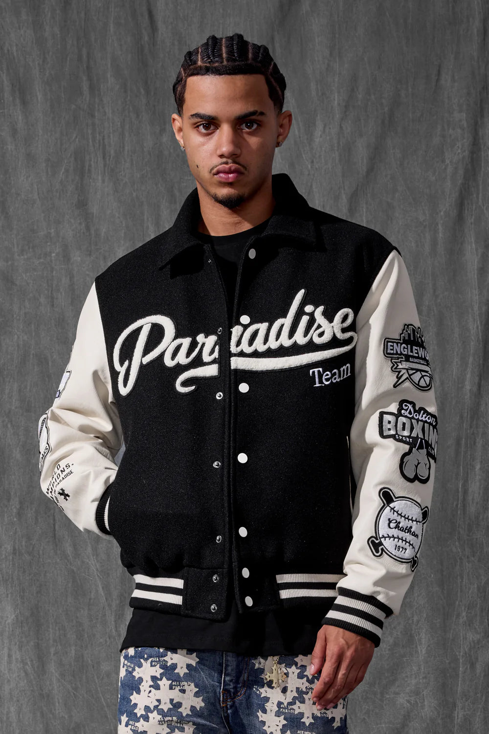 Man wearing a black and white letterman jacket with various logos against a gray background