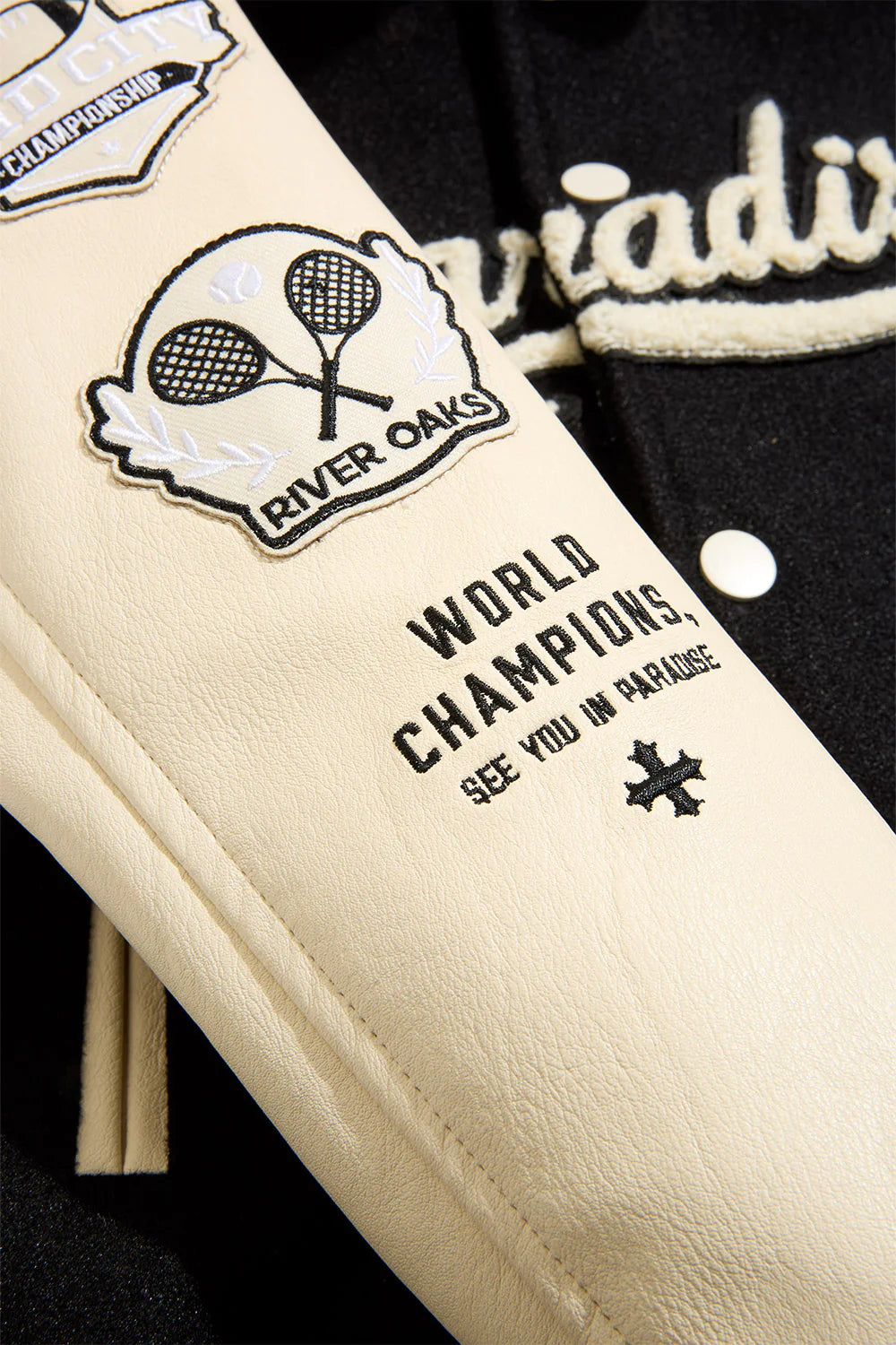 “Paradise Team Varsity Jacket –Black: wool body, quilted satin lining, vegan leather sleeves, chenille patches, embroidered details, and snap button front.”