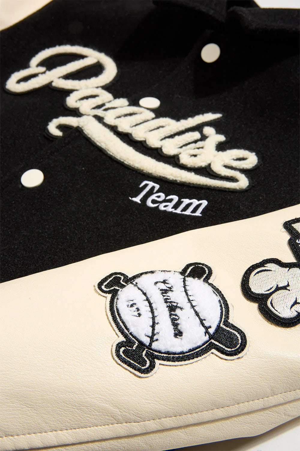 “Paradise Team Varsity Jacket –Black: wool body, quilted satin lining, vegan leather sleeves, chenille patches, embroidered details, and snap button front.”