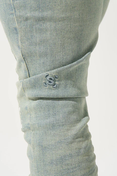 SERENEDE® AURA vintage earth blue jeans with white paint detailing on knee, fitted construction, fading and distressing, leather waistband patch, holographic hangtag, and branded hardware