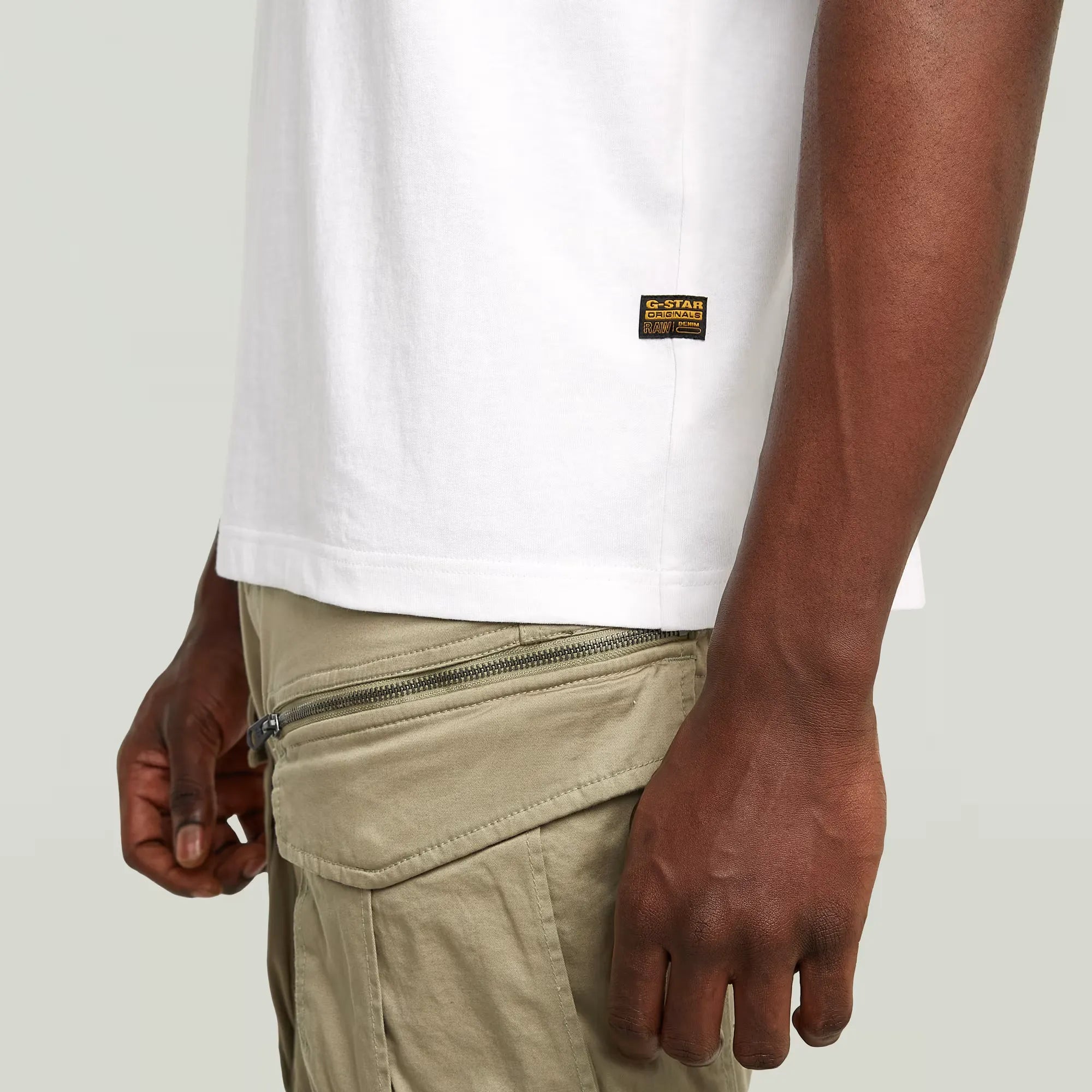 Person wearing a white t-shirt with a brand logo and beige pants on a plain background