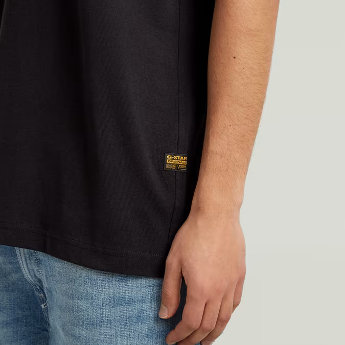 Black t-shirt with a visible brand logo worn by a person, on a plain background