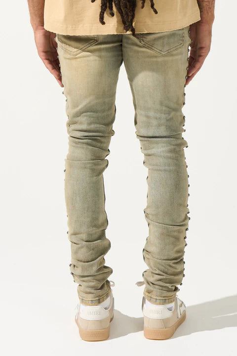 SERENEDE® EXO vintage earth brown jeans with raw sienna overdyed wash, fitted construction, metal studs along stitching, whiskering and fading, leather waistband patch, holographic hangtag, and branded hardware