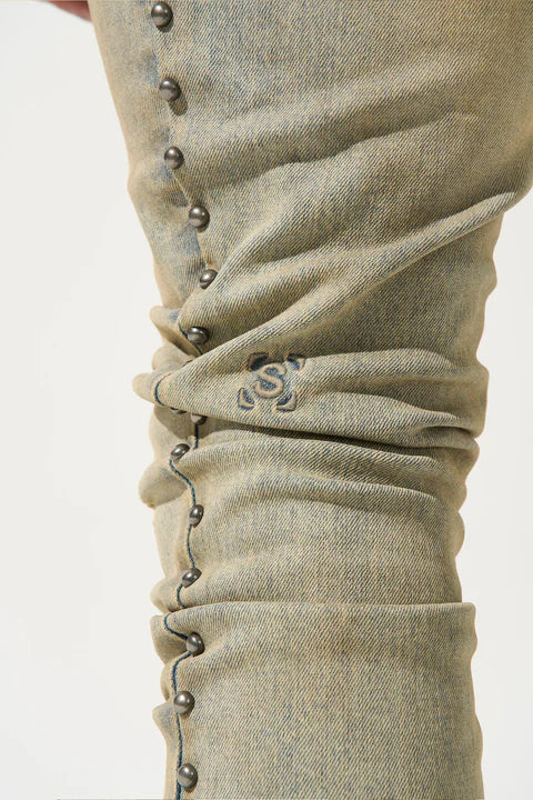 SERENEDE® EXO vintage earth brown jeans with raw sienna overdyed wash, fitted construction, metal studs along stitching, whiskering and fading, leather waistband patch, holographic hangtag, and branded hardware