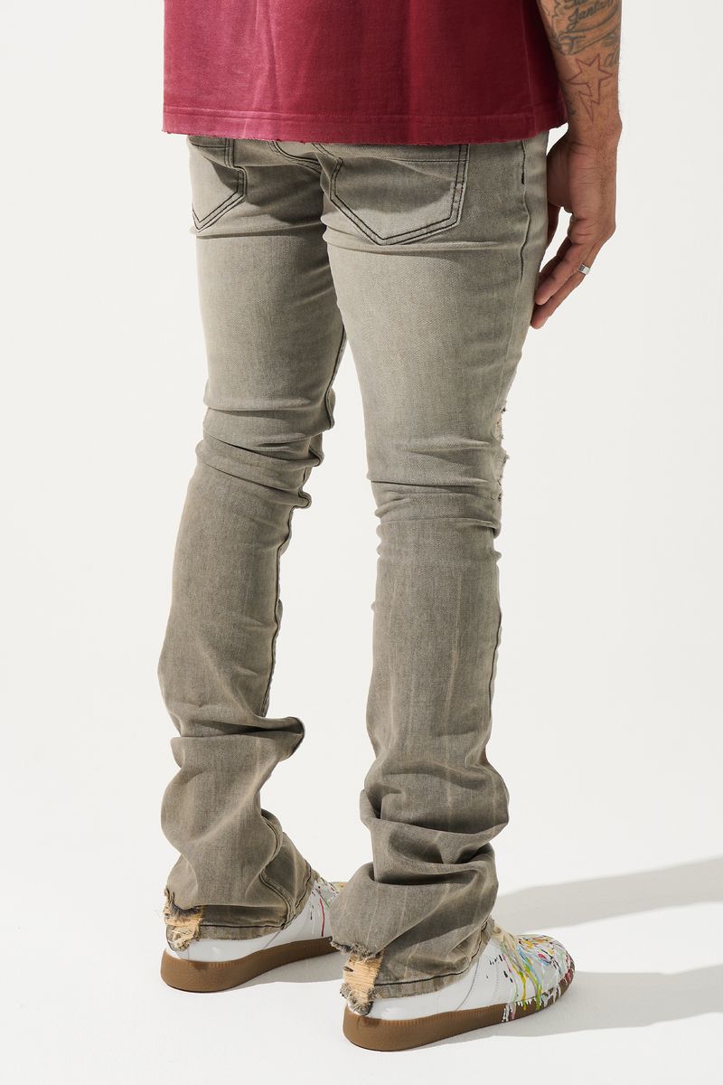 SERENEDE® Nanodust stacked jeans in vintage grey-taupe denim with heavy knee distressing, rust-brown overspray, and extended stacked inseam.