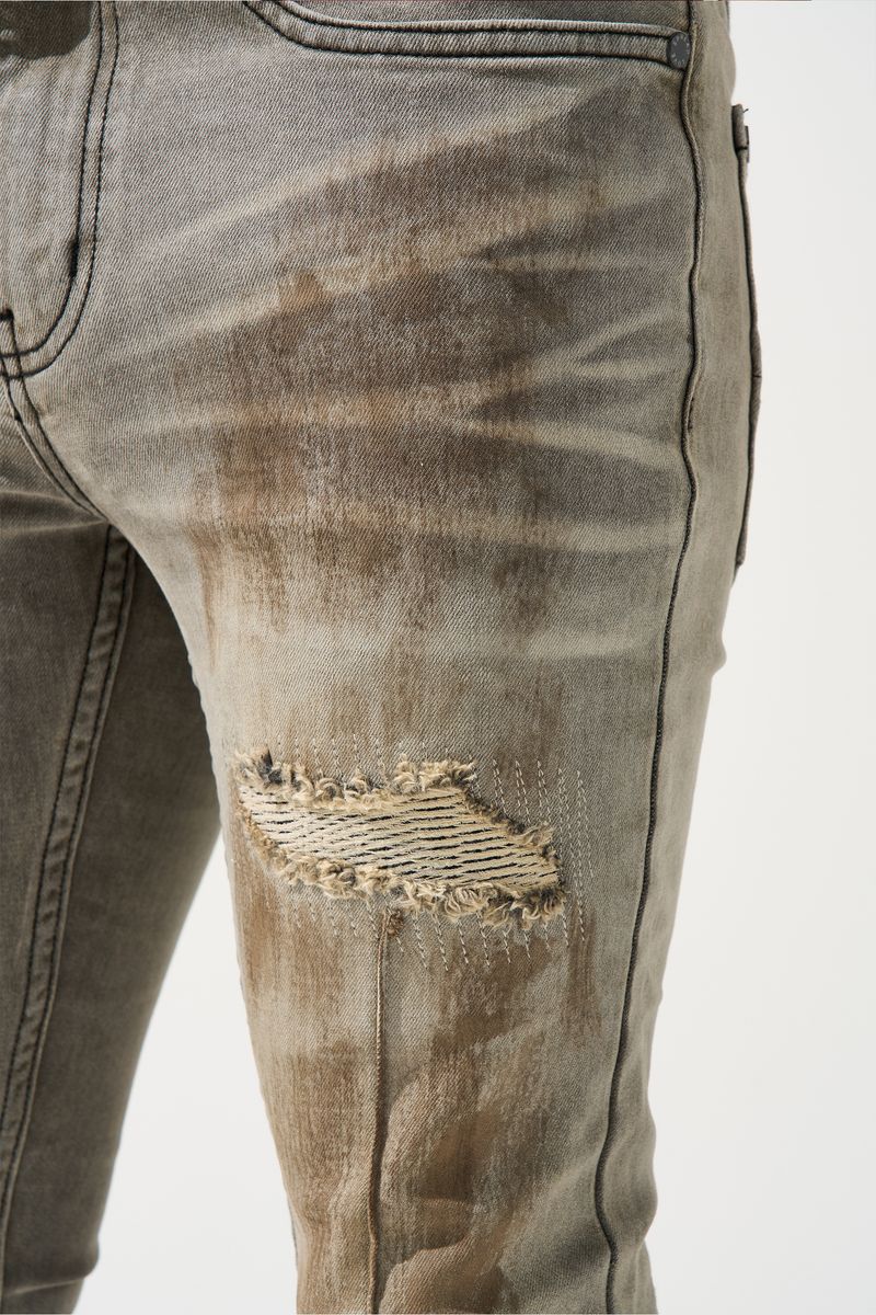 SERENEDE® Nanodust stacked jeans in vintage grey-taupe denim with heavy knee distressing, rust-brown overspray, and extended stacked inseam.