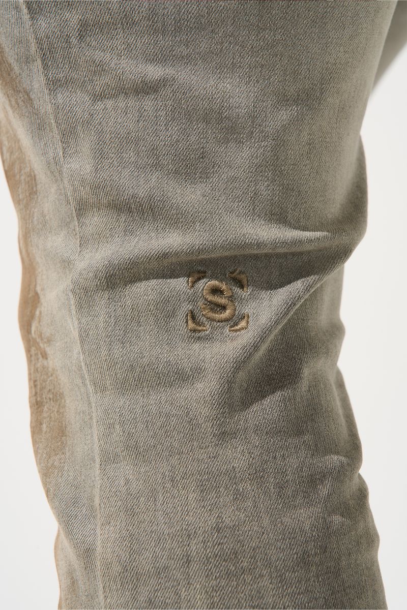 SERENEDE® Nanodust stacked jeans in vintage grey-taupe denim with heavy knee distressing, rust-brown overspray, and extended stacked inseam.